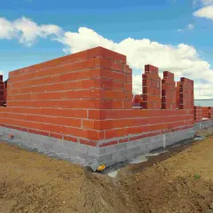 Budowa domu 150m2 w toku. Wznoszone są ściany z czerwonej cegły na betonowym fundamencie. W tle zielone pole i błękitne niebo z chmurami.
