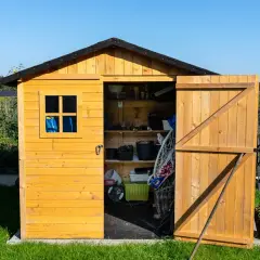 Drewniany domek narzędziowy na trawniku, z otwartymi drzwiami, pełen narzędzi i akcesoriów ogrodowych. Solidne podłoże pod domek narzędziowy zapewnia stabilność.