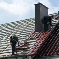 Dwaj dekarze układają dachówki. Widać rusztowanie i komin. Trudno ocenić ile dachówek na m2.