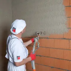 Pracownik w białym ubraniu i czapce z logo Baumit nakłada tynk agregatem na ścianę z pustaków. Obliczasz ile kg tynku na m2?