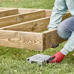 Osoba w rękawiczkach montuje drewnianą konstrukcję, która będzie podstawą dla domku narzędziowego. Podpora z bloczków betonowych czeka na swoje miejsce.