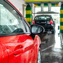 Czerwony samochód w myjni automatycznej, w tle czarny samochód i szczotki myjące.