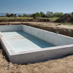 Budowa małego basenu w trakcie. Betonowa niecka w wykopie, otoczona ziemią. W tle domy i drzewa.