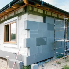 Budowa domu 200m2 w toku. Trwa montaż szarej izolacji na ścianach z białych pustaków. Widać okna i rusztowanie.