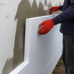 Pracownik w czerwonych rękawicach montuje styropian na elewacji. Zastanawiasz się, ile za metr tynku na elewacji?