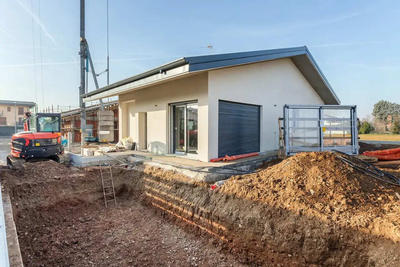 Trwa budowa domku letniskowego. Widoczne są wykopane fundamenty, koparka i materiały budowlane.